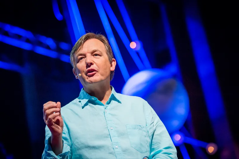 TED の 赤い 円形 ロゴ を 背景 に ステージ で マイク を 持って 話す Chris Anderson 氏